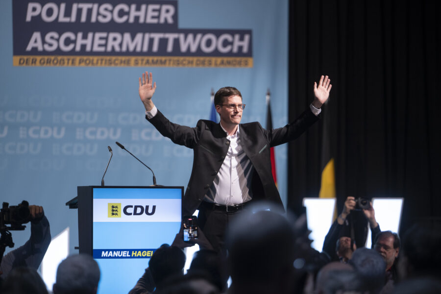 Mann steht auf Bühne mit erhobenen Armen, Podium mit CDU-Logo, Publikum im Vordergrund.