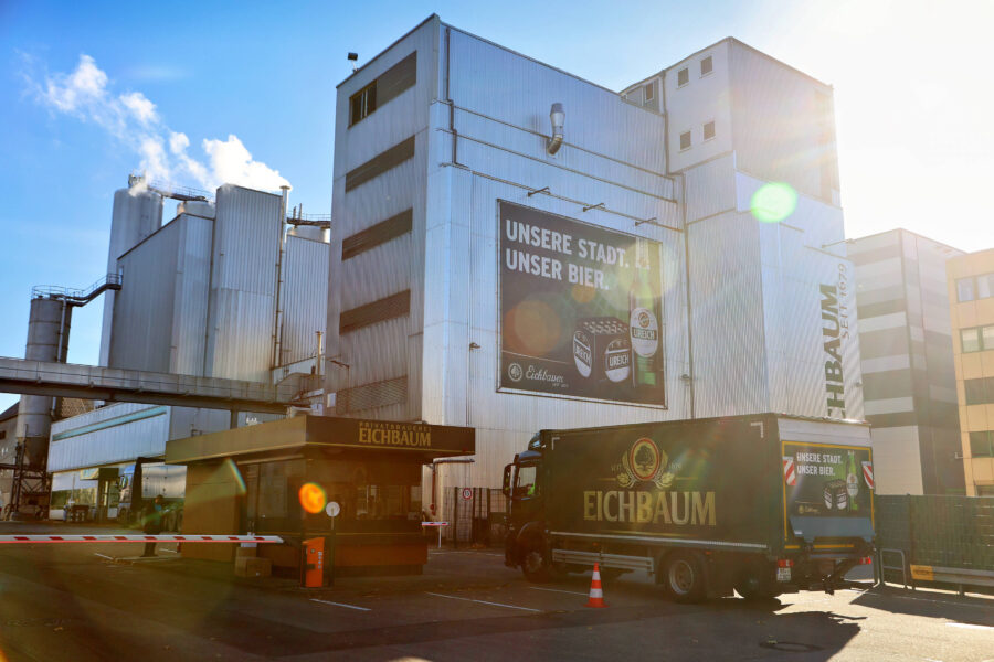Industriegebäude mit "Eichbaum"-Logo, Lastwagen und Werbeschild im Sonnenlicht.