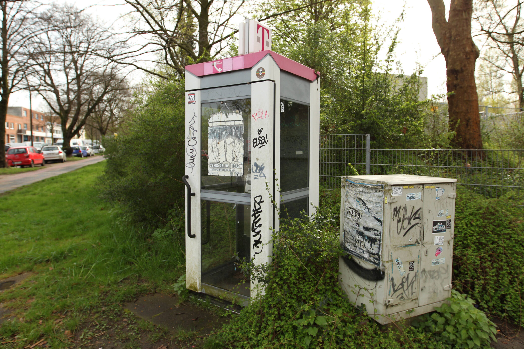 Telefonzelle mit Graffiti, daneben ein Kasten, umgeben von Bäumen und Gras.