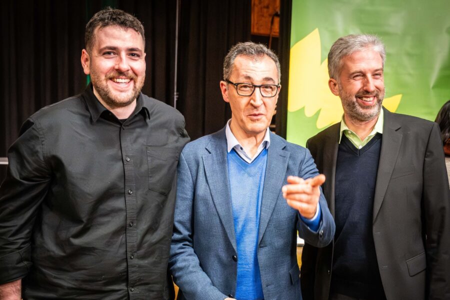 Drei lachende Männer in Anzügen stehen nebeneinander vor einem grünen Hintergrund.