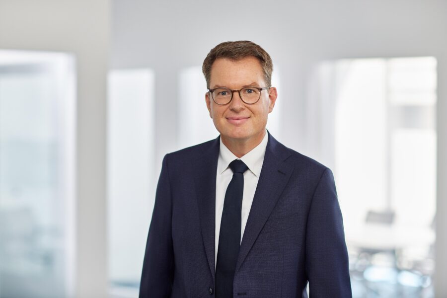 Mann in Anzug mit Brille, in einem hellen Raum stehend.