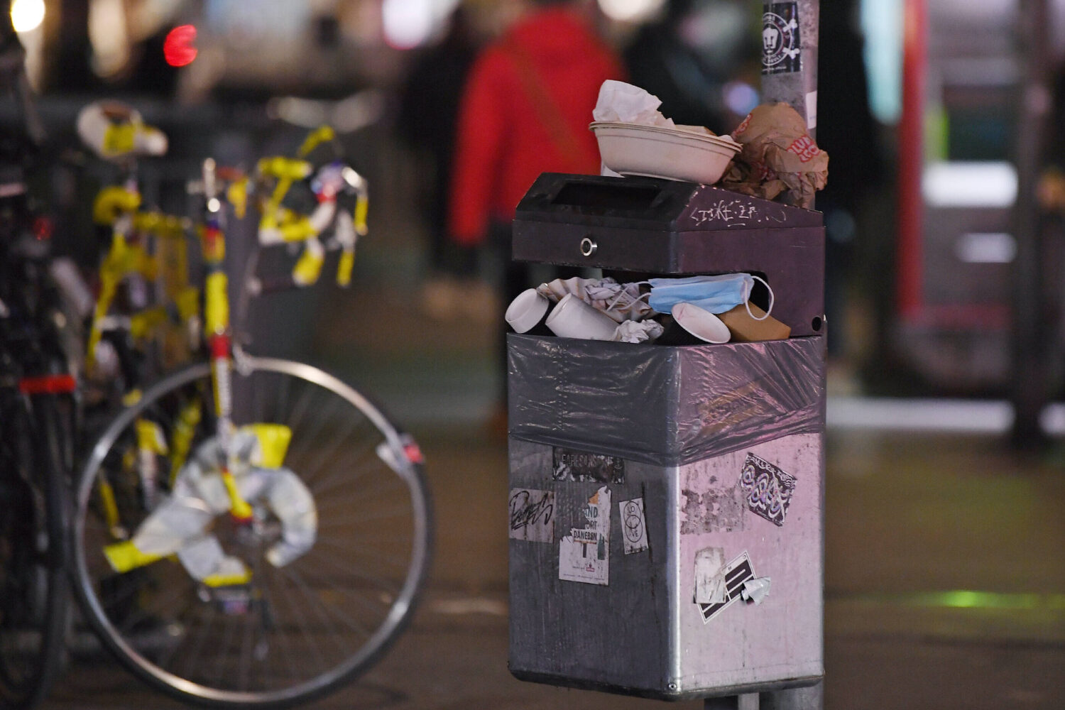Überfüllter Mülleimer mit Masken und Bechern, Fahrrad im Hintergrund.