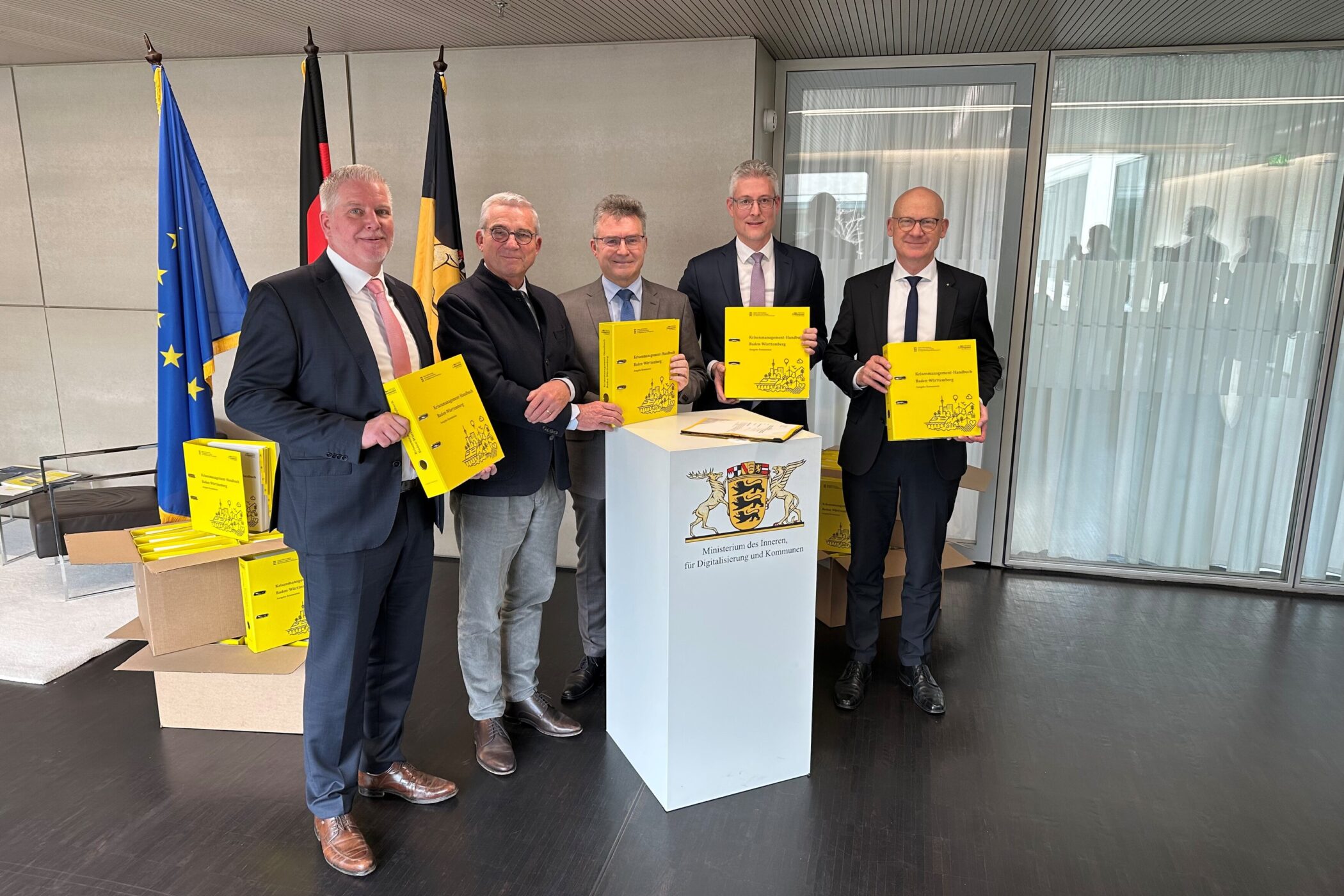 Fünf Männer in Anzügen halten gelbe Broschüren, im Hintergrund Flaggen.