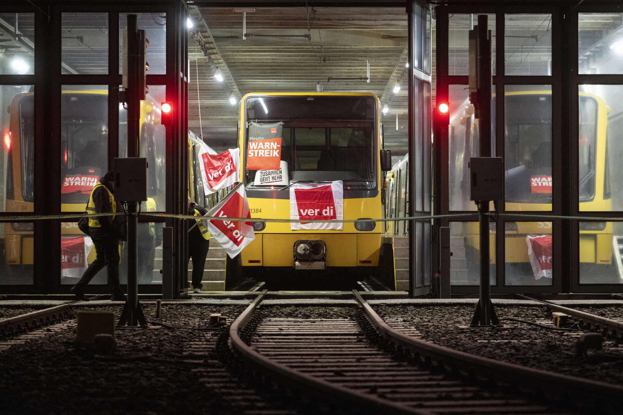 Zwei gelbe Züge im Depot mit Streikschildern und ver.di-Flaggen, zwei Personen in Warnwesten.
