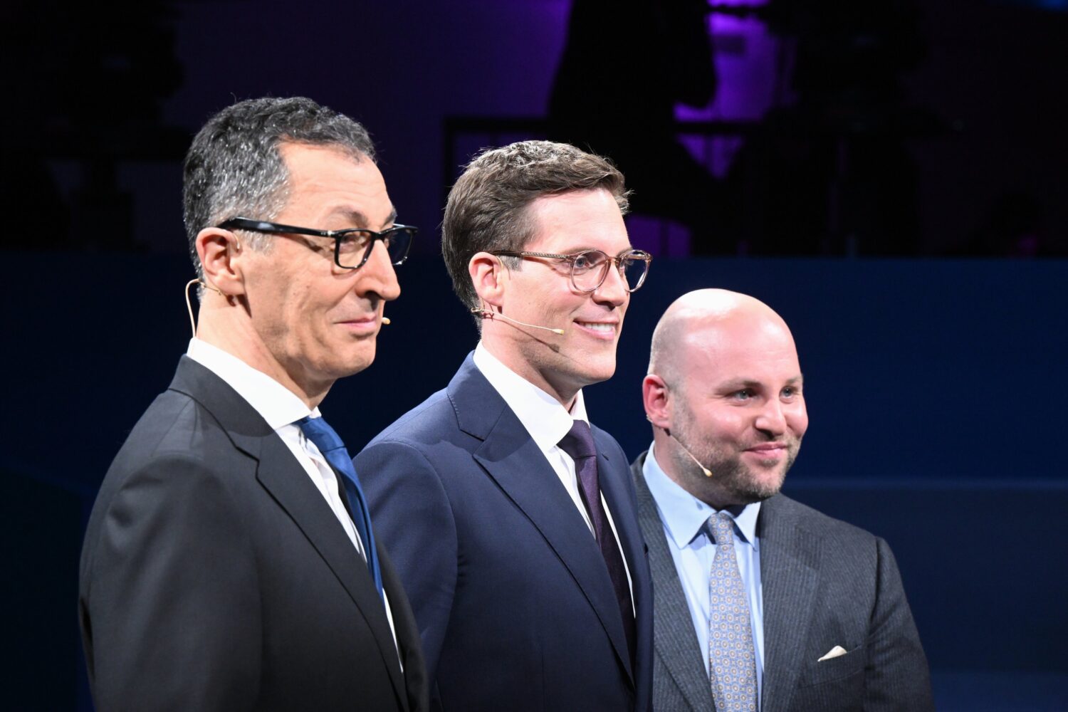 Drei Männer in Anzügen mit Headsets stehen nebeneinander.