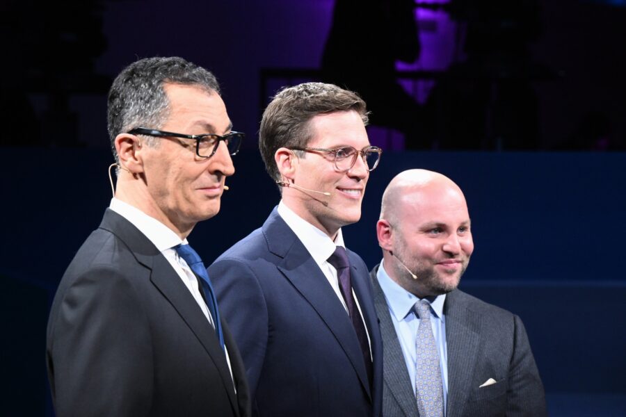 Drei Männer in Anzügen mit Headsets stehen nebeneinander.