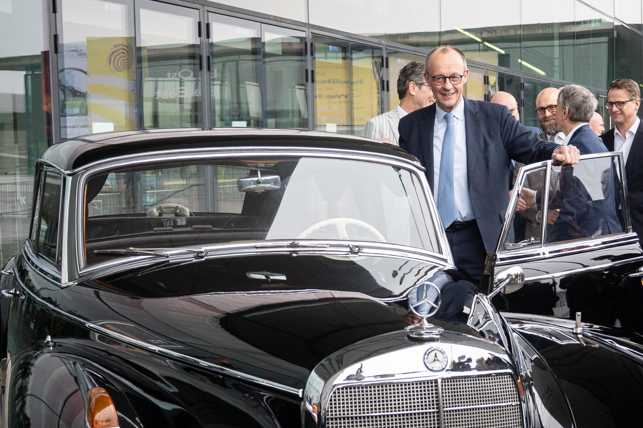 Sieben Männer um ein schwarzes Mercedes-Oldtimer-Cabrio vor einem Gebäude.