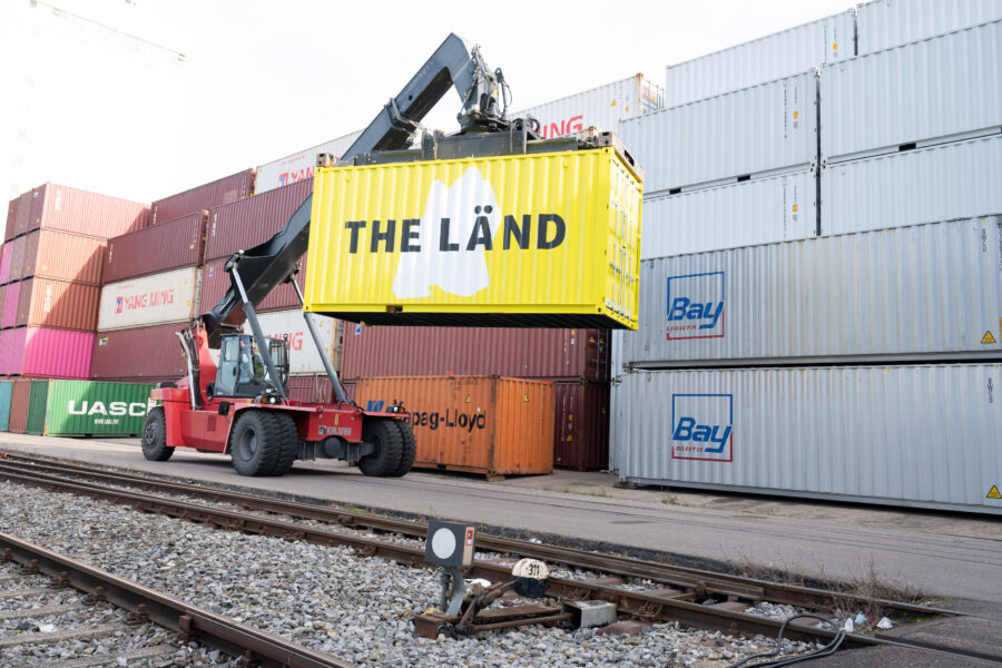 Gelber Container mit "THE LÄND" wird von rotem Kranfahrzeug angehoben.