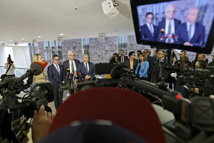 Menschen halten eine Pressekonferenz, Kameras filmen, Zuschauer stehen dabei.