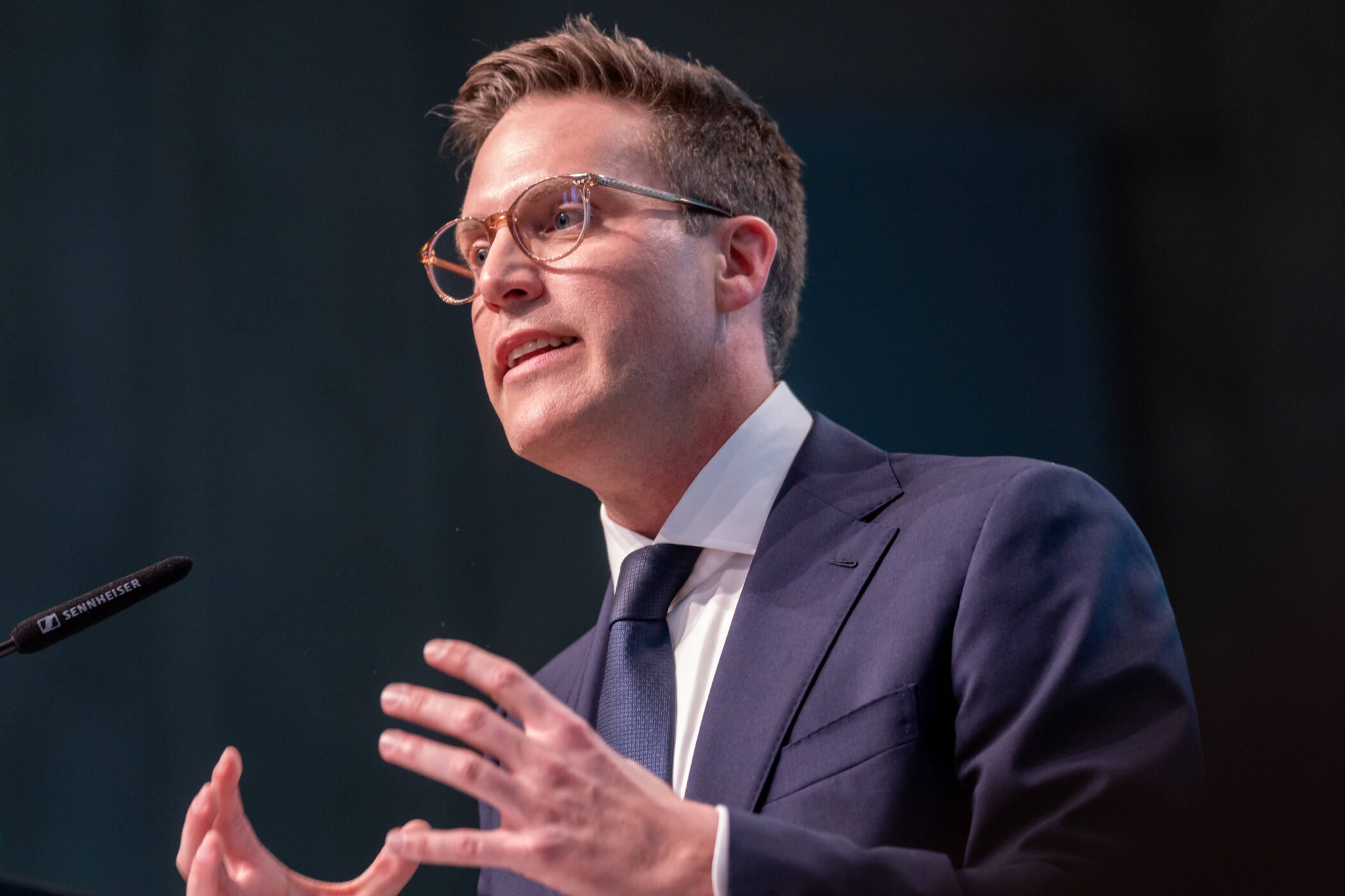 Mann in Anzug und Brille spricht am Mikrofon.