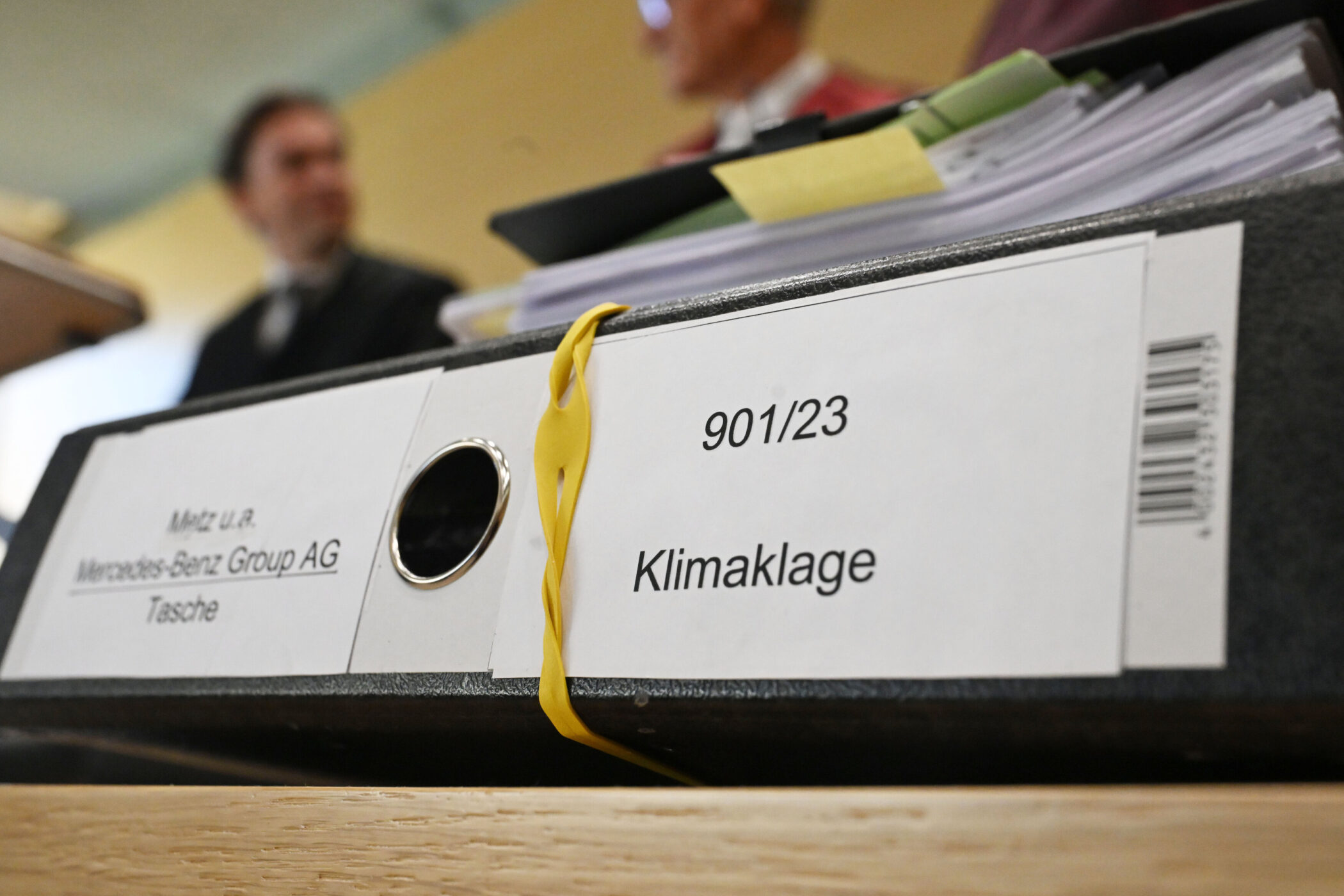 Aktenordner mit Aufschrift "Klimaklage 901/23", zwei unscharfe Personen im Hintergrund.