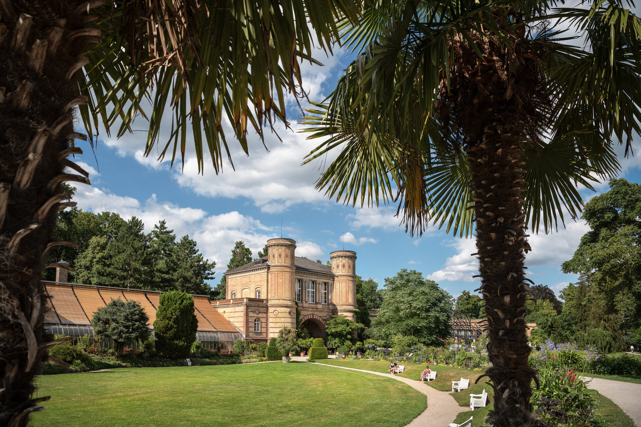 Backsteingebäude mit Türmen, Park, Palmen, Bänke, Spaziergänger, blauer Himmel.