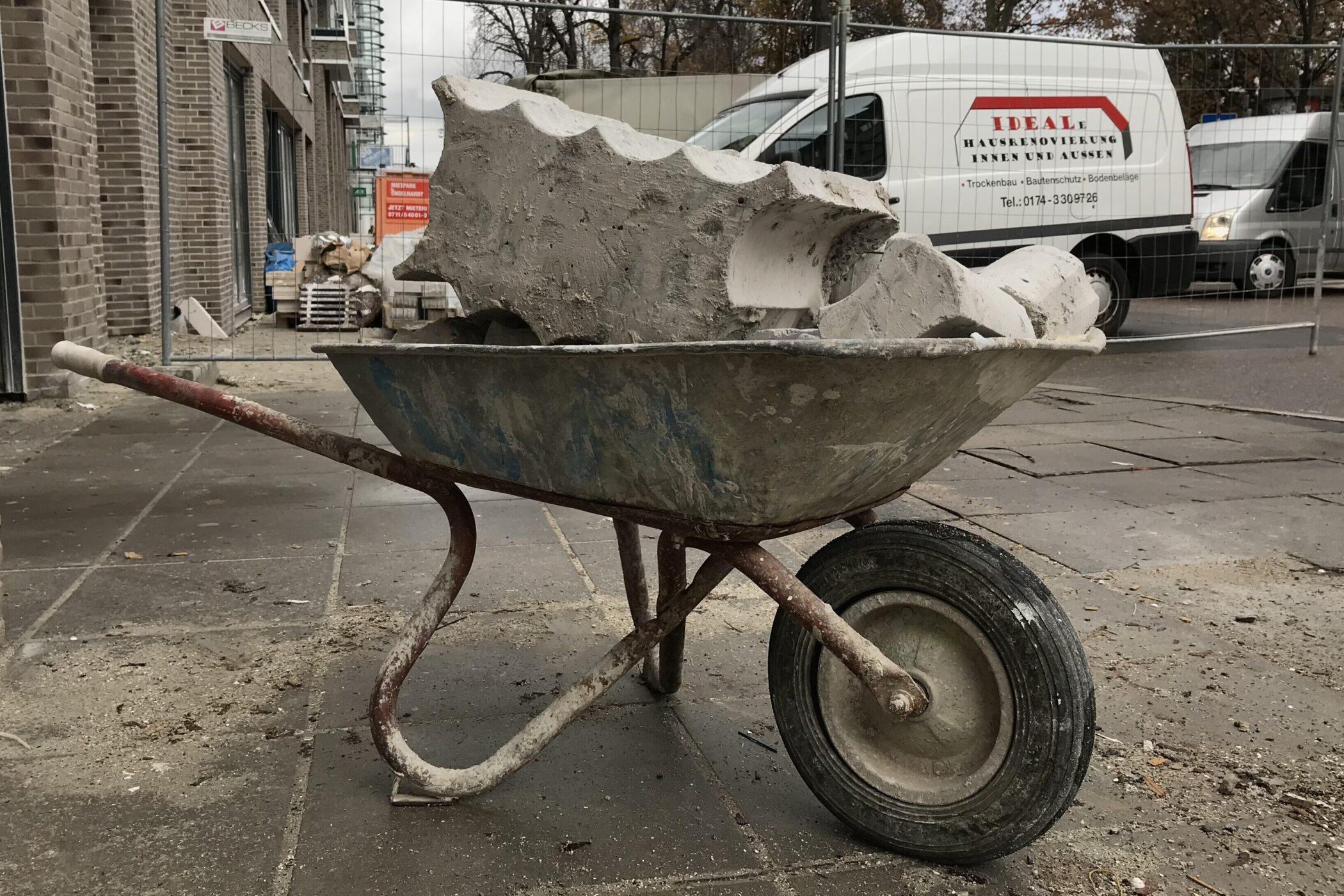 Schubkarre mit Schutt vor Baustelle, parkender Lieferwagen im Hintergrund.