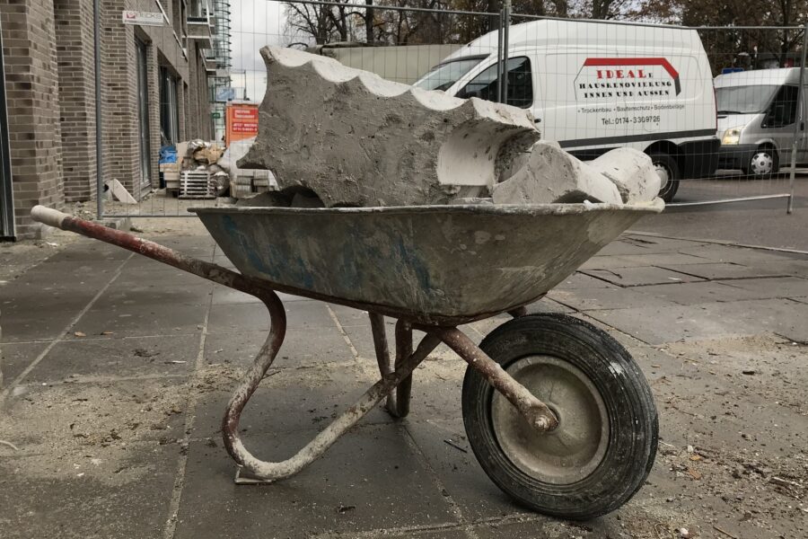 Schubkarre mit Schutt vor Baustelle, parkender Lieferwagen im Hintergrund.