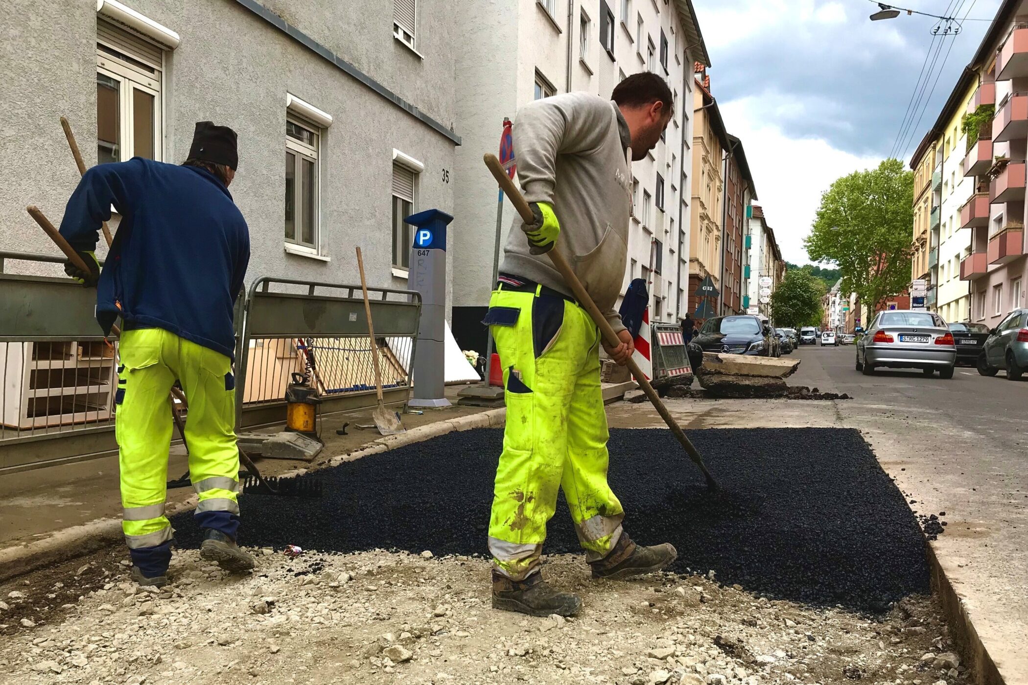 Zwei Arbeiter asphaltieren eine Straße in einer Wohngegend.