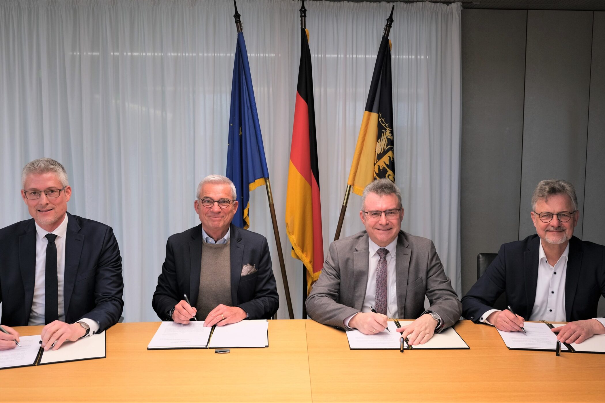Vier Männer sitzen an einem Tisch und unterschreiben Dokumente. Im Hintergrund Fahnen.