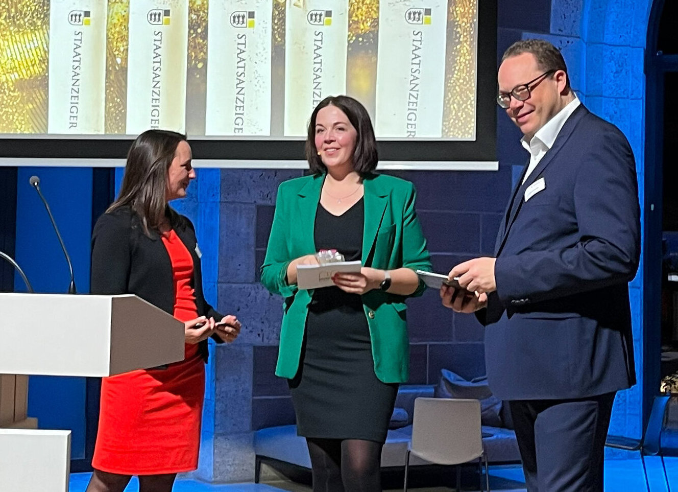 Drei Personen in Businesskleidung auf einer Bühne mit Podium und Bildschirm.