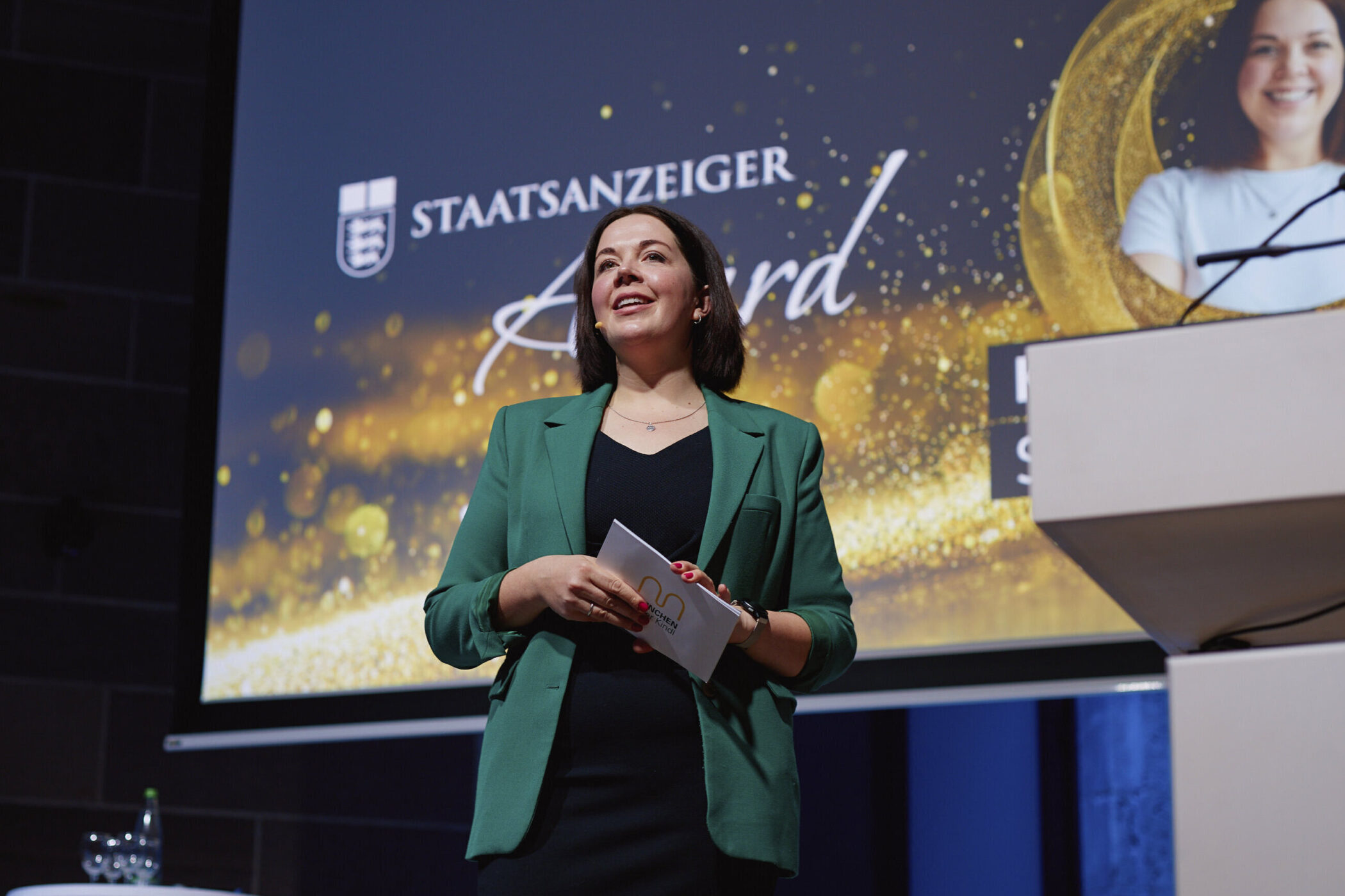 Frau in grünem Blazer hält Sprechkarten, "STAATSANZEIGER Award" im Hintergrund.