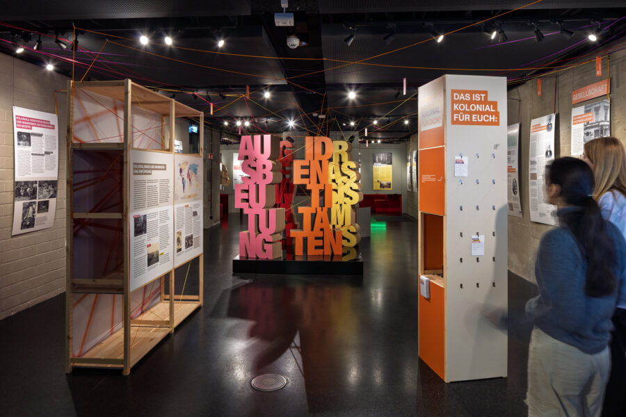 Innenraum mit Ausstellungstafeln, Wandtexten, Holzrahmen und farbigen Buchstaben.