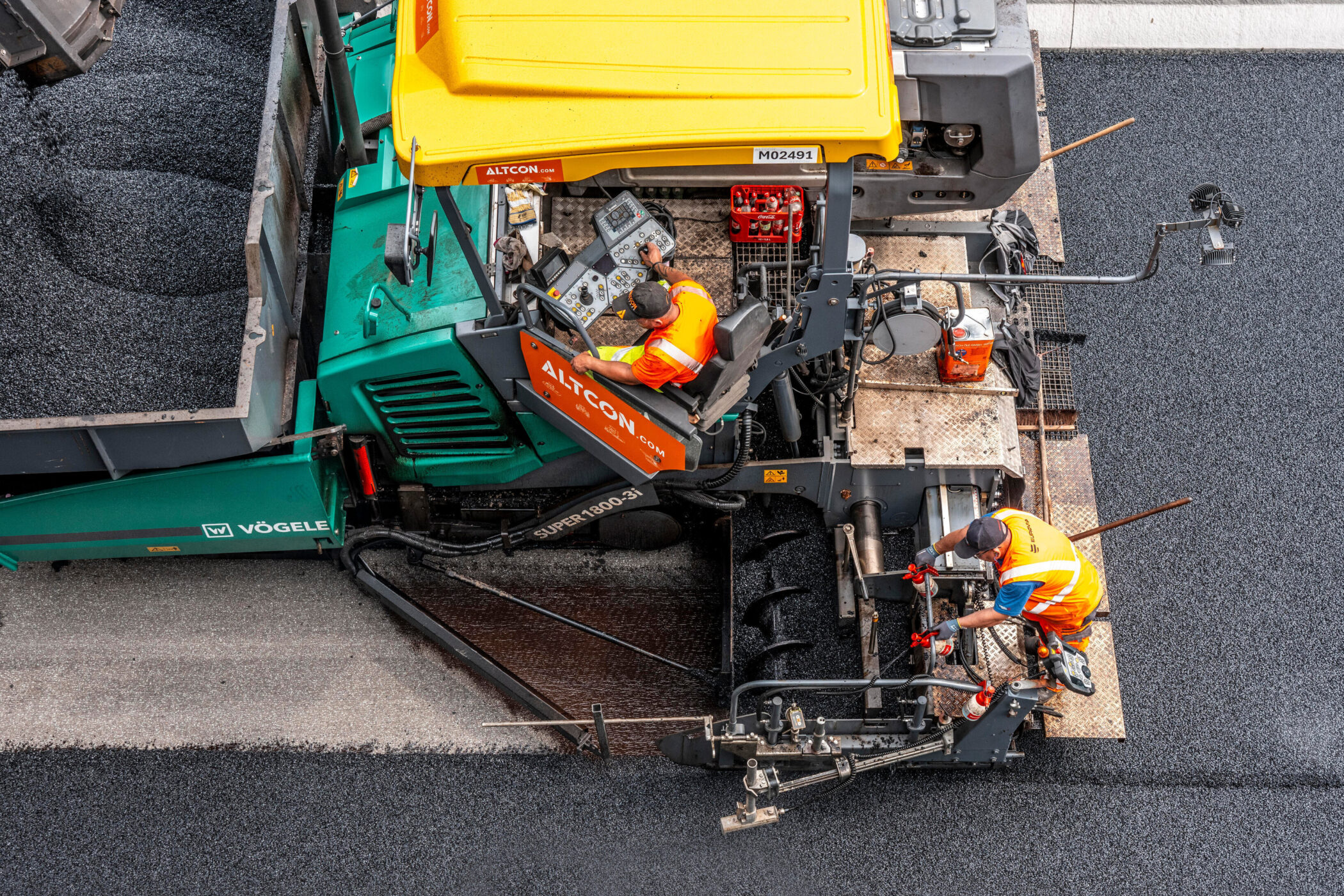Zwei Arbeiter in orange Westen bedienen Asphaltfertiger auf einer Straße.