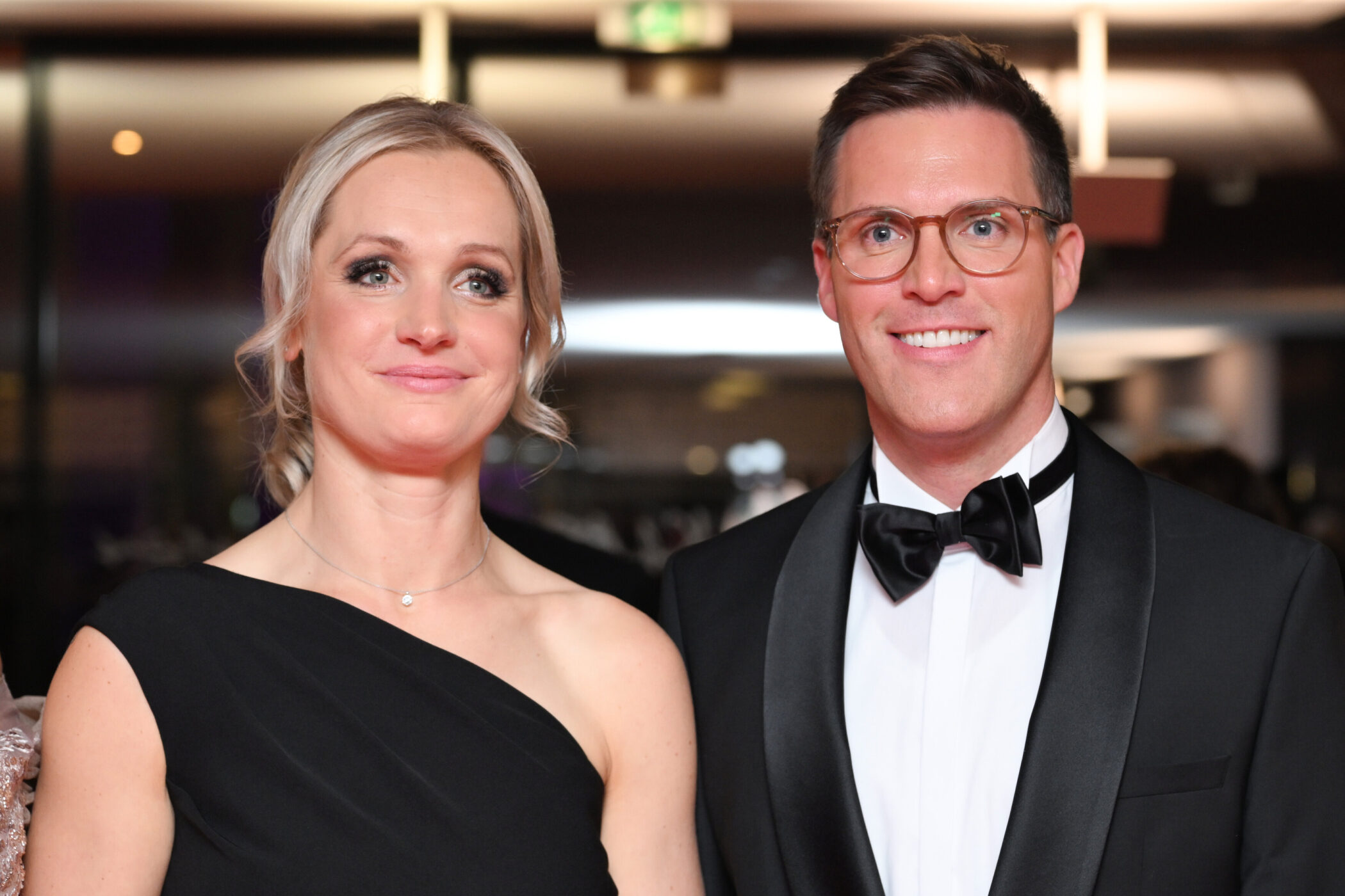 Zwei lächelnde Personen in Abendkleidung vor unscharfem Hintergrund.