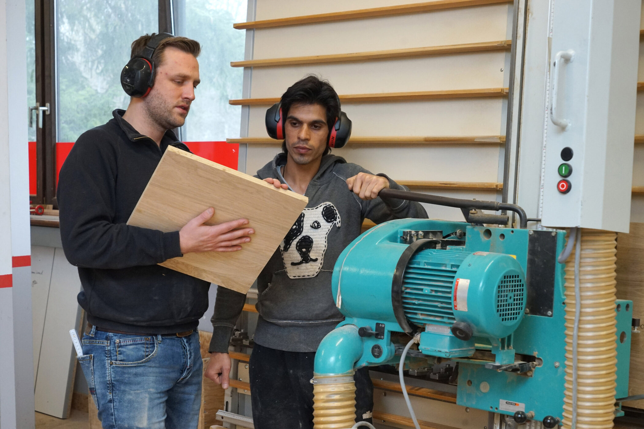 Zwei Männer mit Gehörschutz an einer Maschinenanlage. Einer hält ein Holzbrett.