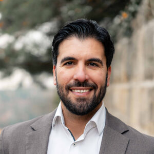 Ein lächelnder Mann in einem Anzug vor unscharfem Hintergrund.