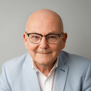 Ein lächelnder Mann mit Brille in einem hellblauen Sakko vor grauem Hintergrund.