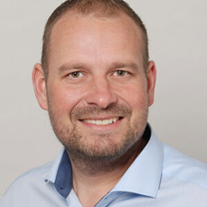 Mann mit Hemd lächelt in die Kamera. Hintergrund ist einfarbig hell.