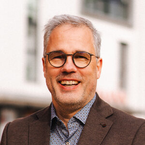 Ein lächelnder Mann mit Brille und Sakko im Freien.