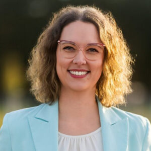 Eine Person mit Brille lächelt, trägt ein helles Jackett und steht im Freien.