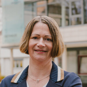 Eine Frau mit kurzem Haar lächelt in die Kamera, Gebäude im Hintergrund.