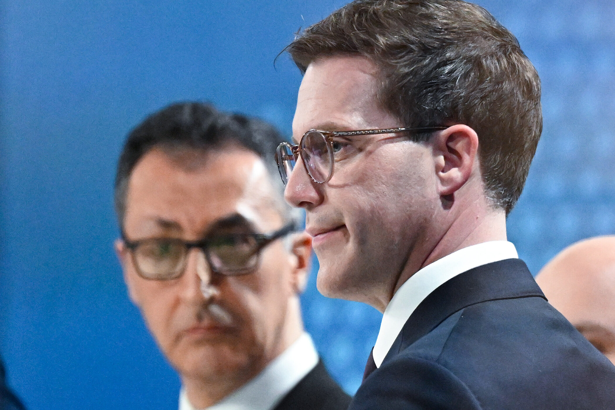 Zwei Männer mit Brillen, seitlich nebeneinander, unscharfer blauer Hintergrund.