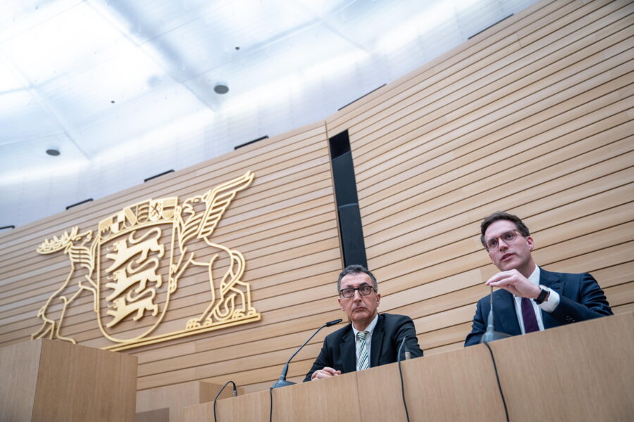 Zwei Männer sitzen hinter einem Rednerpult vor einem Wandwappen.