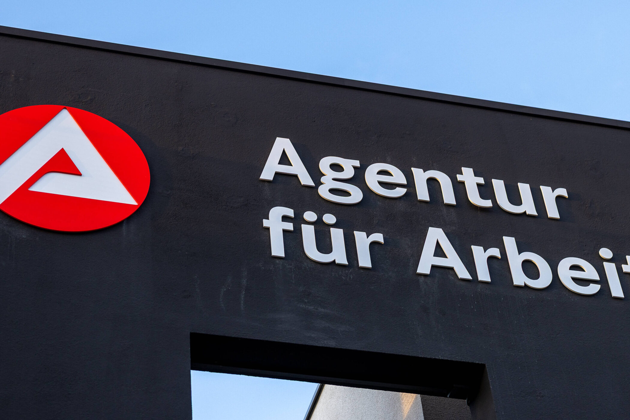 Logo der Agentur für Arbeit auf dunklem Gebäude, blauer Himmel.
