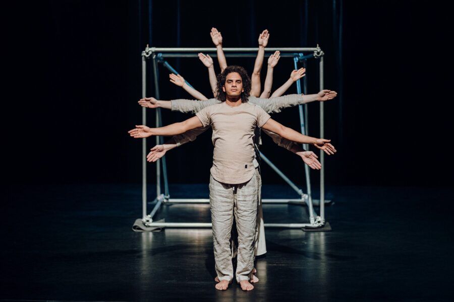 Zwei Personen, eine mit vielen ausgestreckten Armen dahinter, auf einer dunklen Bühne.