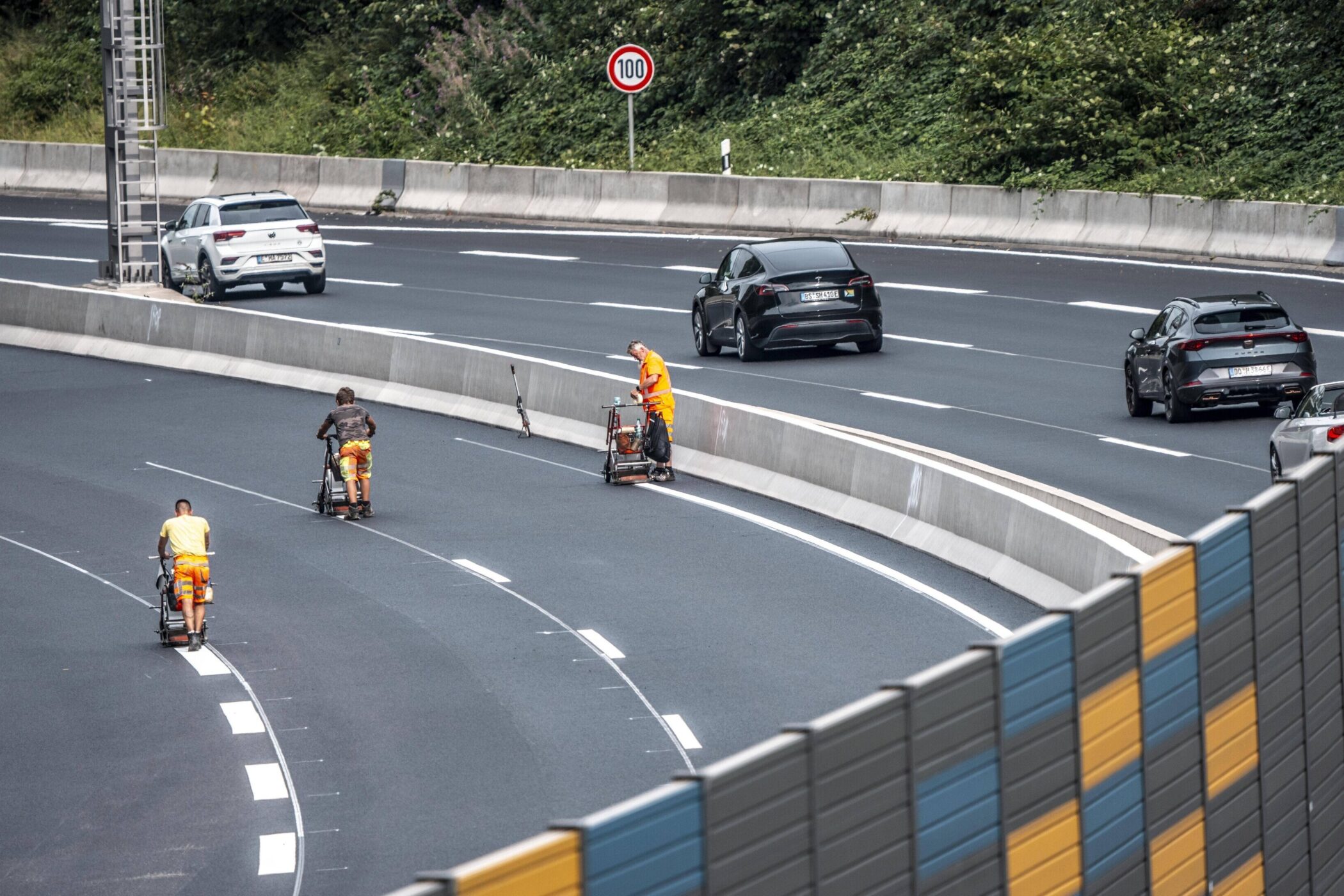Drei Personen markieren Fahrbahnlinien auf Autobahn; Autos fahren vorbei.