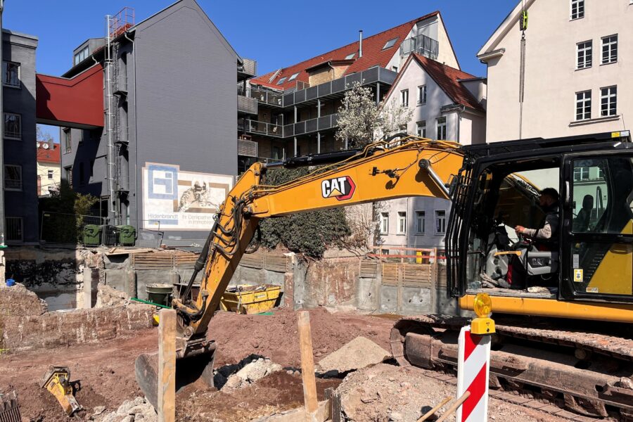 Bagger auf Baustelle, umgeben von Wohnhäusern, eine Person im Führerhaus.