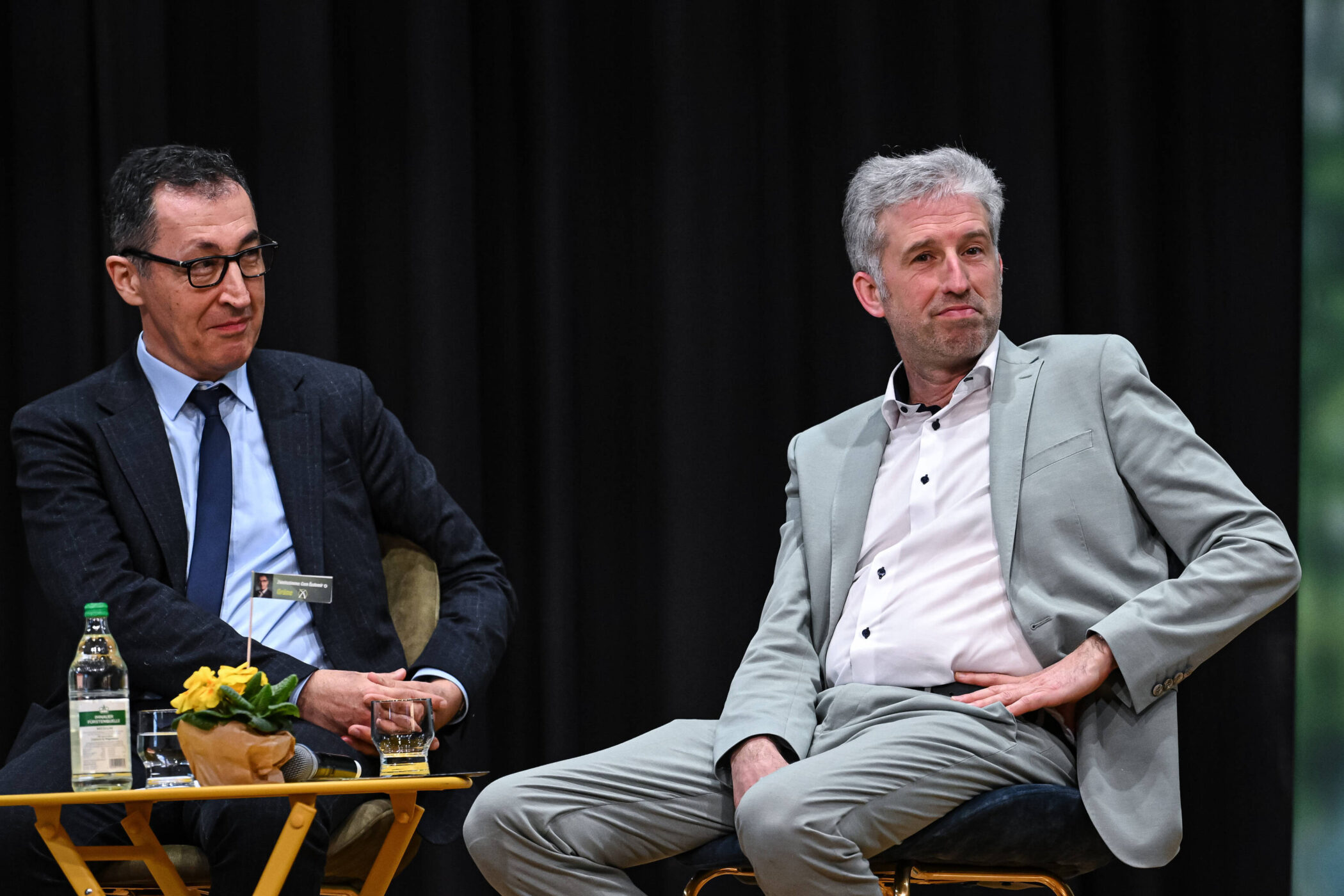 Zwei Männer sitzen auf Stühlen, ein Tisch mit Flaschen und Pflanze vor ihnen.