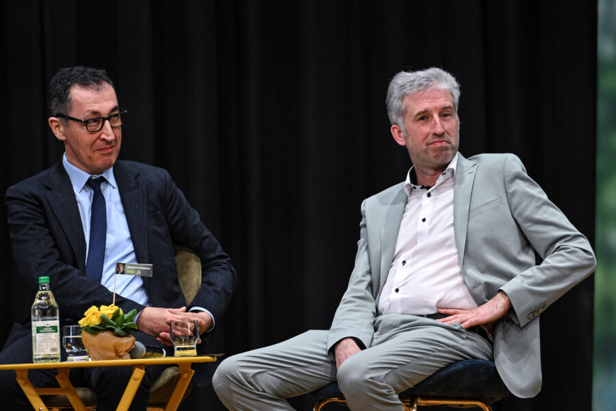 Zwei Männer sitzen auf Stühlen, ein Tisch mit Flaschen und Pflanze vor ihnen.