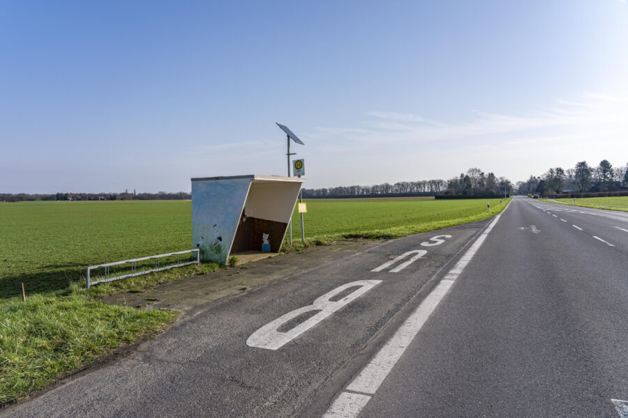 Bushaltestelle mit Überdachung und Solarzelle an einer Landstraße, grüne Felder daneben.