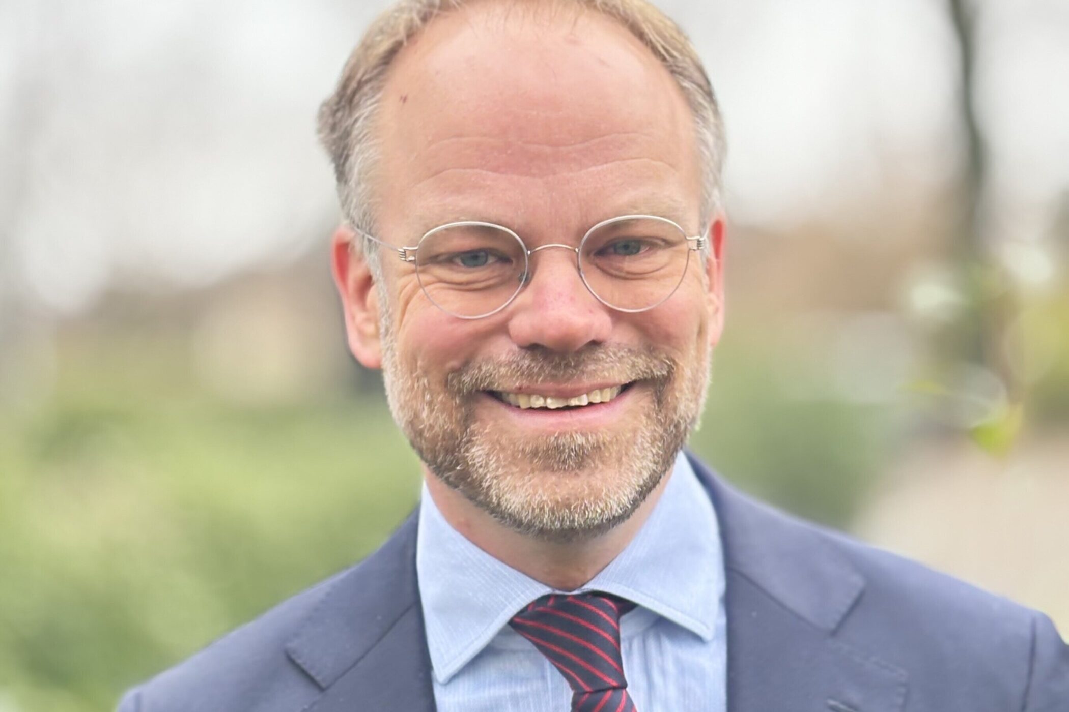 Ein lächelnder Mann mit Brille und Anzug im Freien.