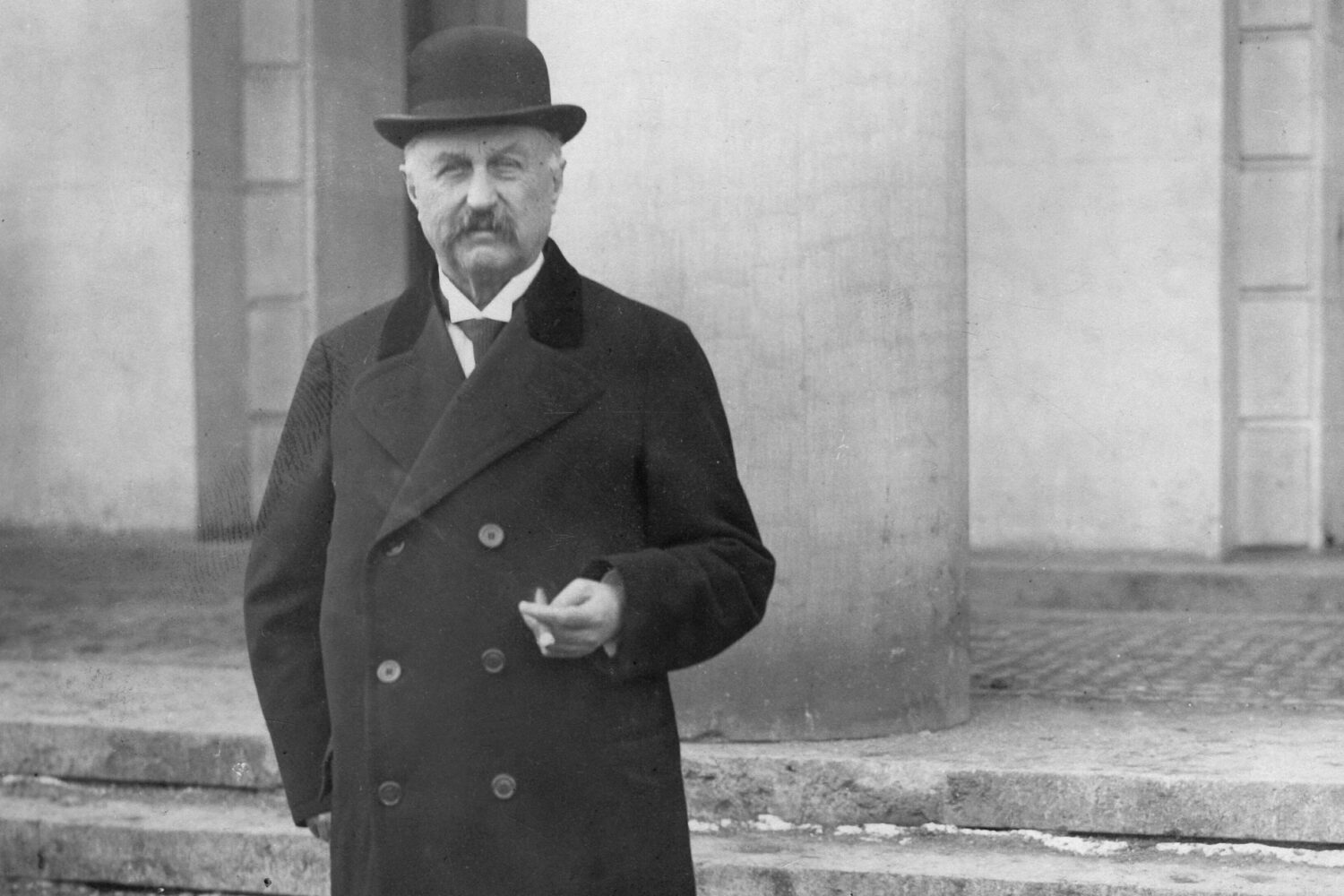 Älterer Mann mit Hut und Mantel steht vor einer Säule.