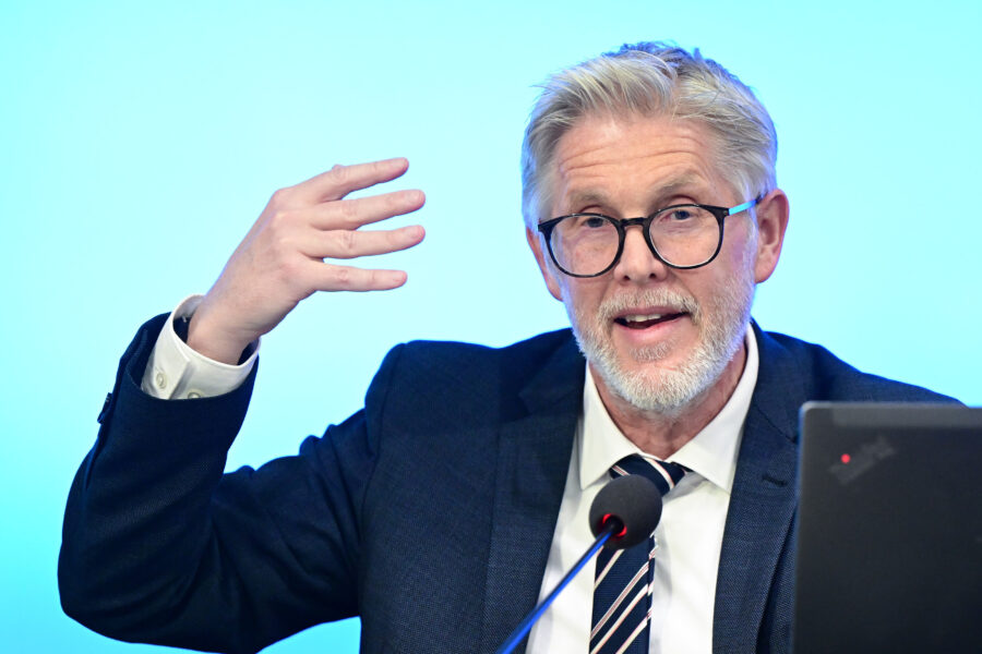 Person mit Brille spricht in ein Mikrofon, blaue Hintergrundbeleuchtung.