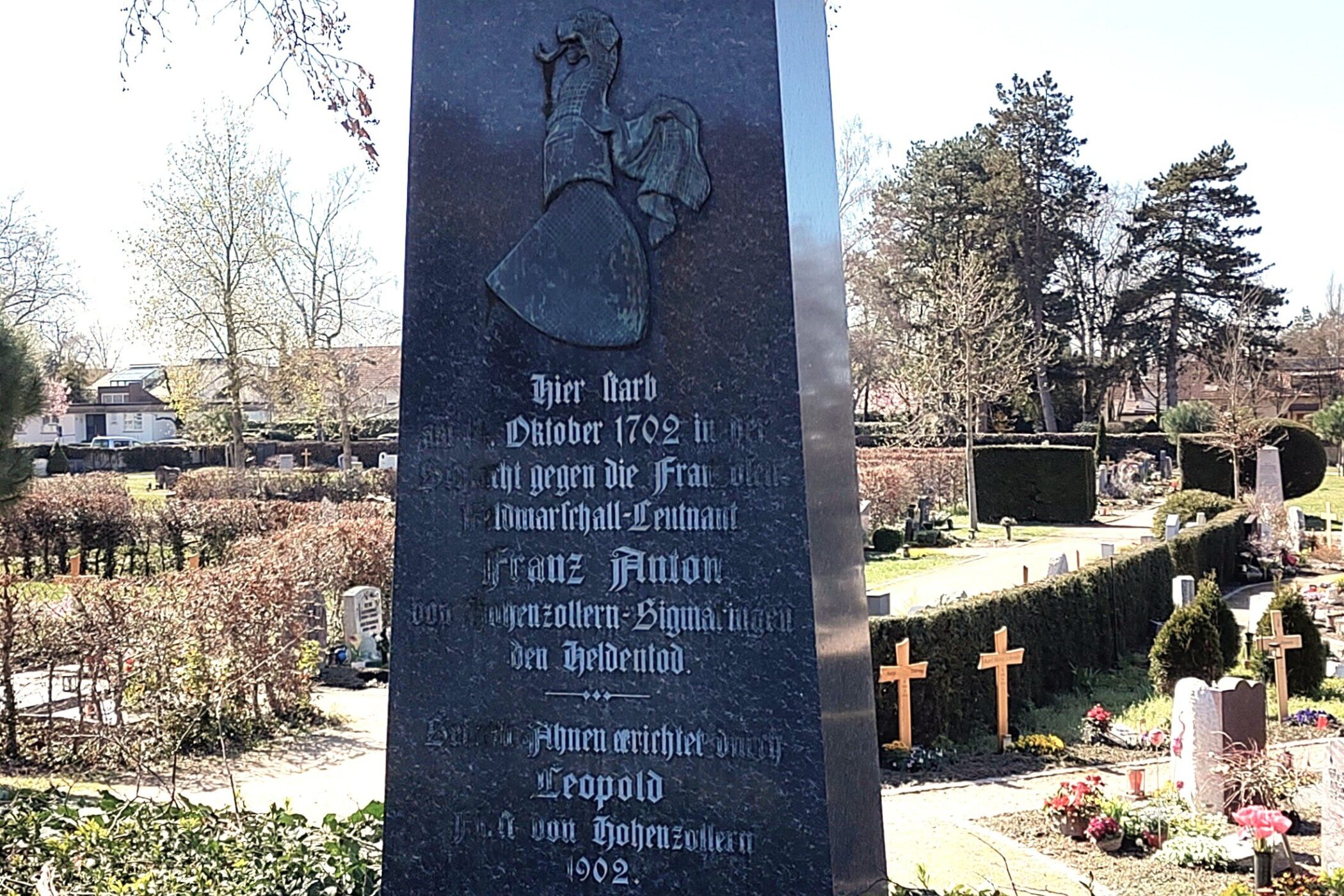 Dunkler Grabstein mit Inschrift; Grabfeld im Hintergrund. Bäume und blauer Himmel.