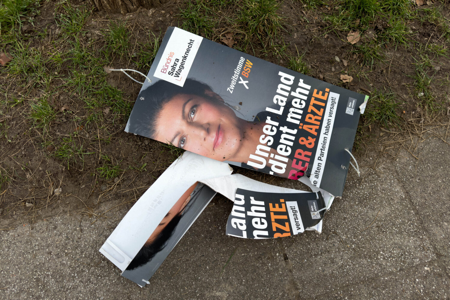 Zerbrochenes Wahlplakat auf dem Boden, teilweise auf Gras und Gehweg.