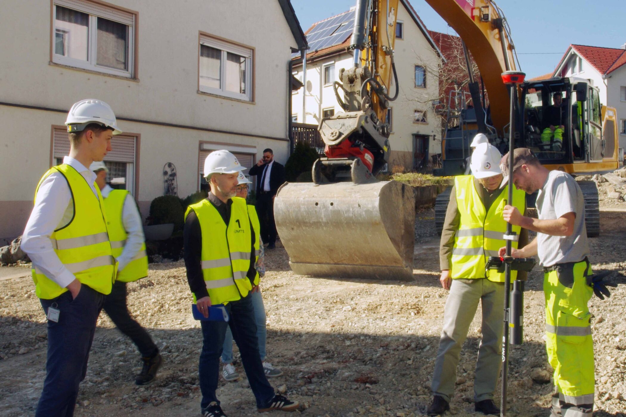 Fünf Personen mit Warnwesten und Helmen auf einer Baustelle vor einem Bagger.