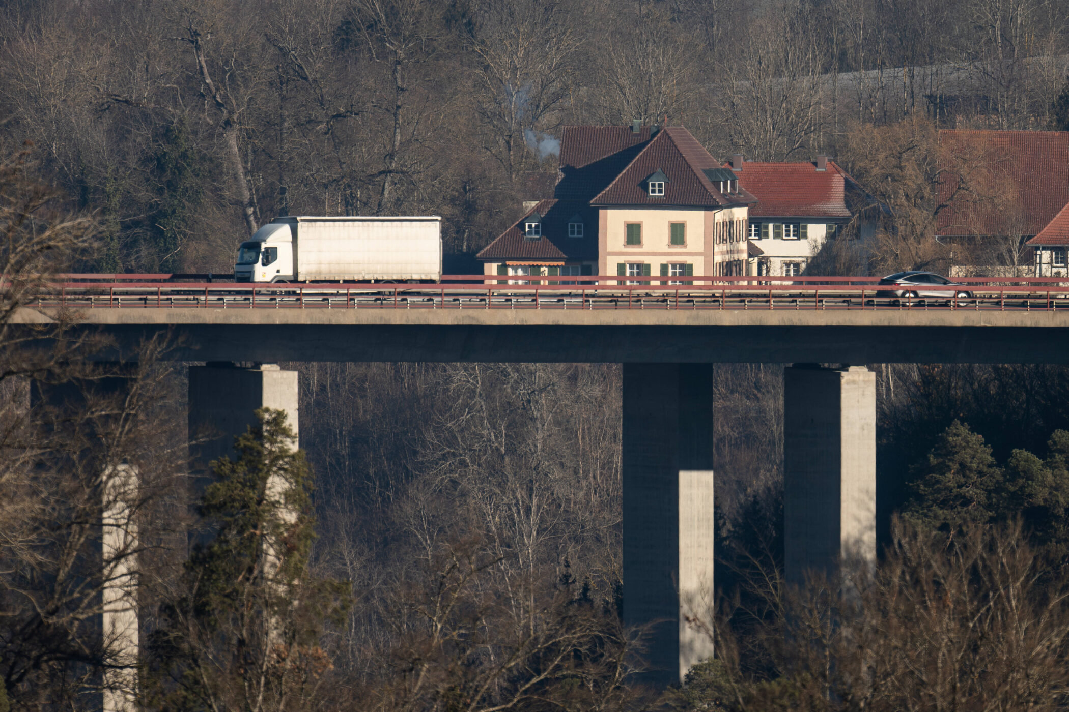 LKW und Auto fahren auf Brücke vor mehreren Häusern, unbelaubte Bäume im Hintergrund.
