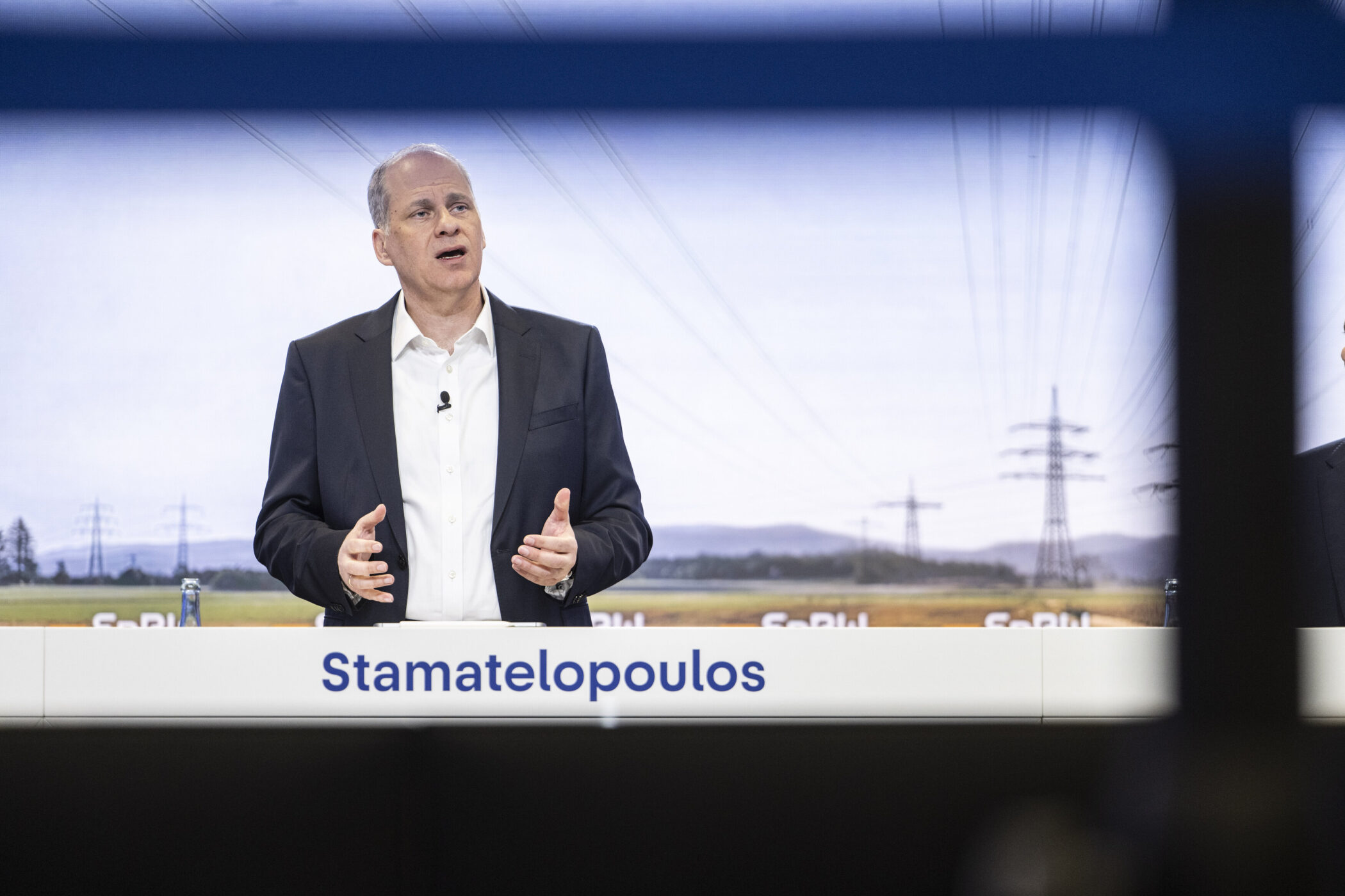 Mann in Anzug vor Landschaftsbild, Name "Stamatelopoulos" auf einem Schild.