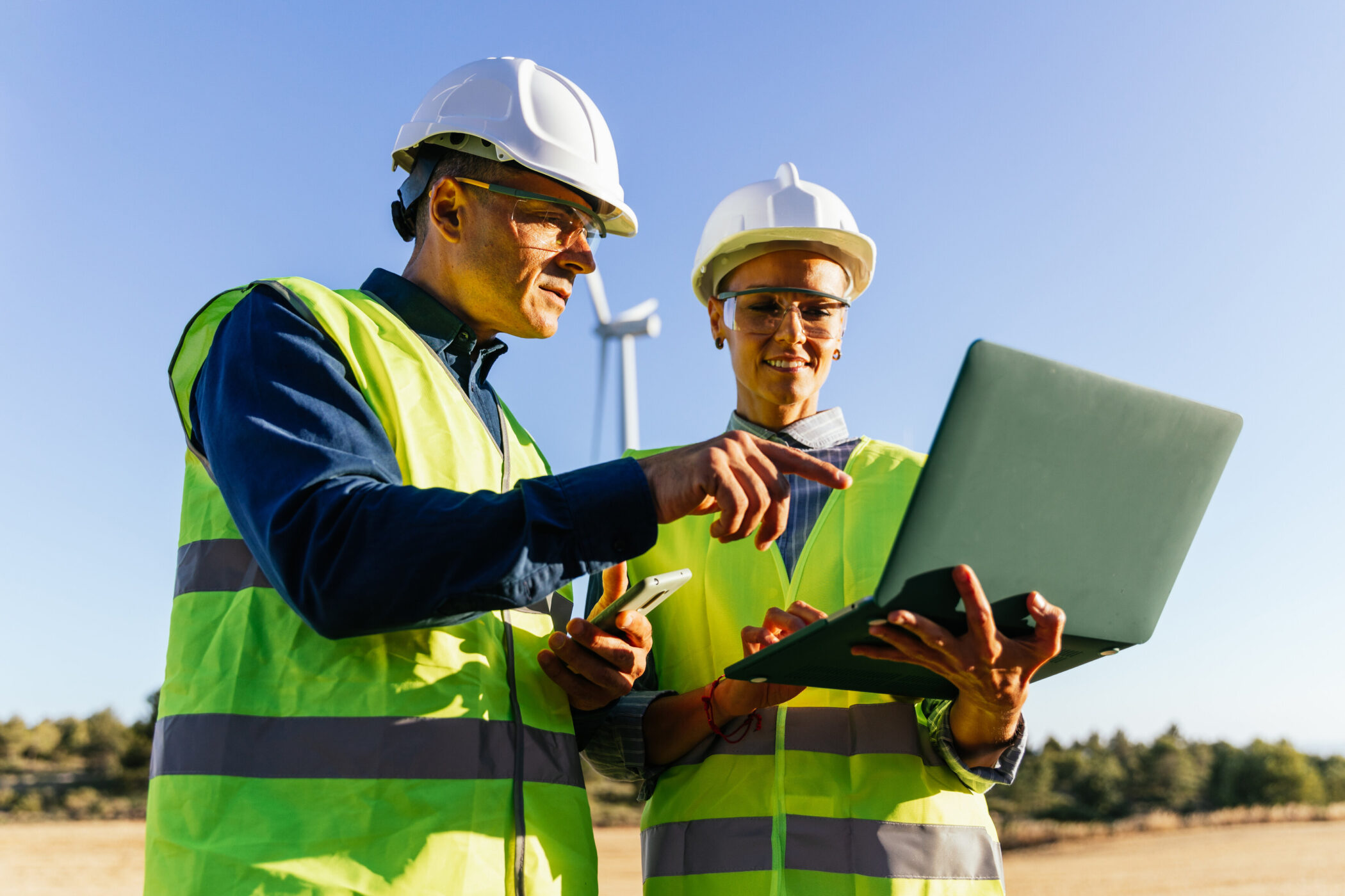 Zwei Personen in Schutzhelmen und Westen mit Laptop im Freien.